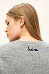 Close-up of the back of a gray knit sweater with the phrase ‘but nice’ embroidered in small navy blue script near the neckline. The person’s straight, light brown hair is draped over the back, and they are wearing small gold hoop earrings.