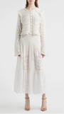 A person wearing a white long‑sleeve blouse and matching midi skirt made from eyelet lace fabric. The blouse features a round neckline, decorative buttons down the front, ruffled shoulders, and flared sleeves with embroidered cutout patterns. The skirt has a smocked waistband and a tiered design with coordinating eyelet embroidery throughout. The outfit is paired with nude heeled sandals. The background is plain and light-colored.
