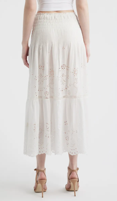 Back view of a person wearing a white long‑sleeve blouse and matching midi skirt made from eyelet lace fabric. The blouse features a round neckline, decorative buttons down the front, ruffled shoulders, and flared sleeves with embroidered cutout patterns. The skirt has a smocked waistband and a tiered design with coordinating eyelet embroidery throughout. The outfit is paired with nude heeled sandals. The background is plain and light-colored.
