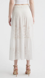 Back view of a person wearing a white long‑sleeve blouse and matching midi skirt made from eyelet lace fabric. The blouse features a round neckline, decorative buttons down the front, ruffled shoulders, and flared sleeves with embroidered cutout patterns. The skirt has a smocked waistband and a tiered design with coordinating eyelet embroidery throughout. The outfit is paired with nude heeled sandals. The background is plain and light-colored.