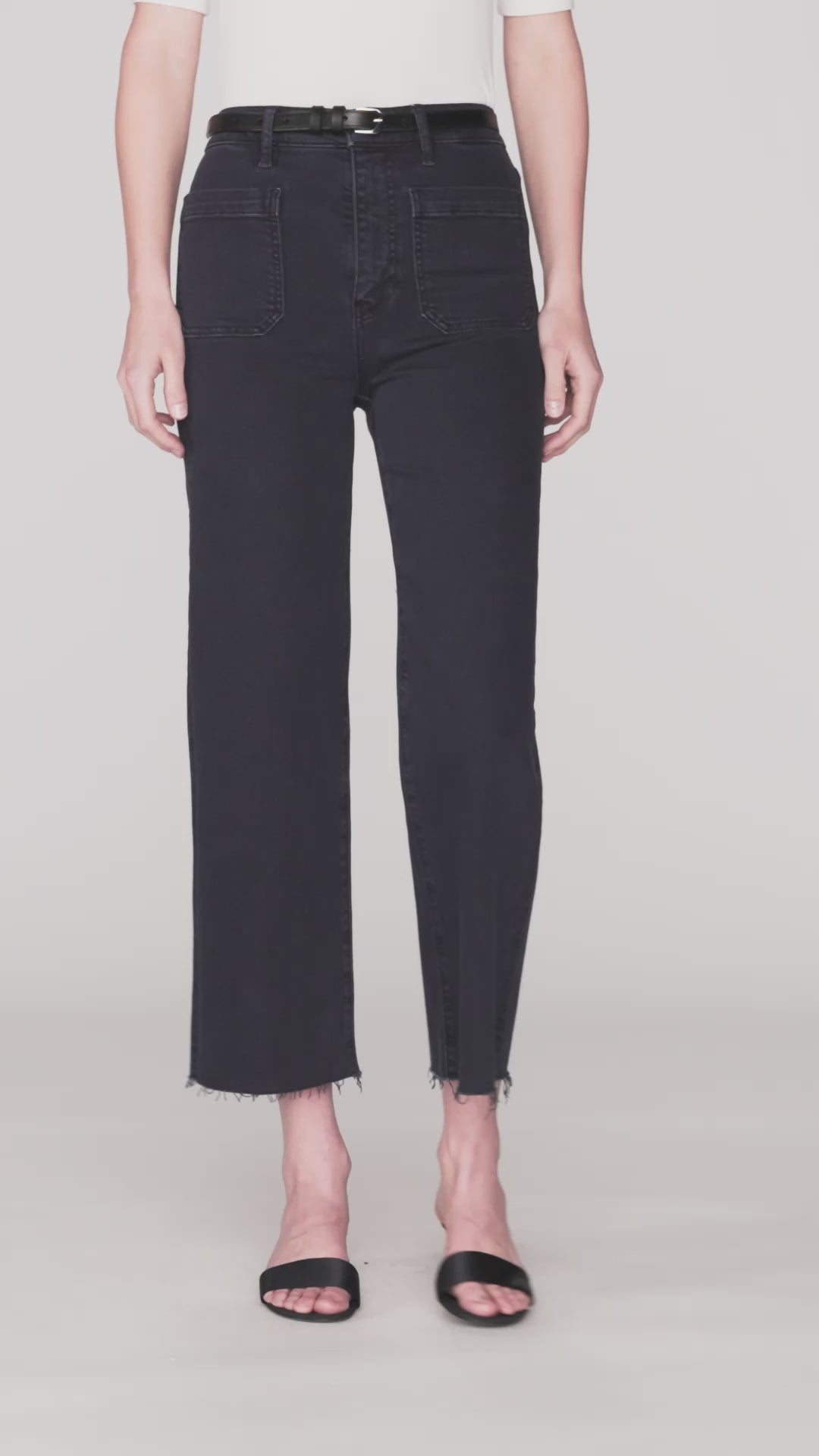 A short clip of a person wearing a white short-sleeve top tucked into high-waisted, navy denim jeans with a black belt. The jeans have zipper and button closure, two large front and back pockets, and frayed hems at the bottom. The person is also wearing black open-toe sandals.