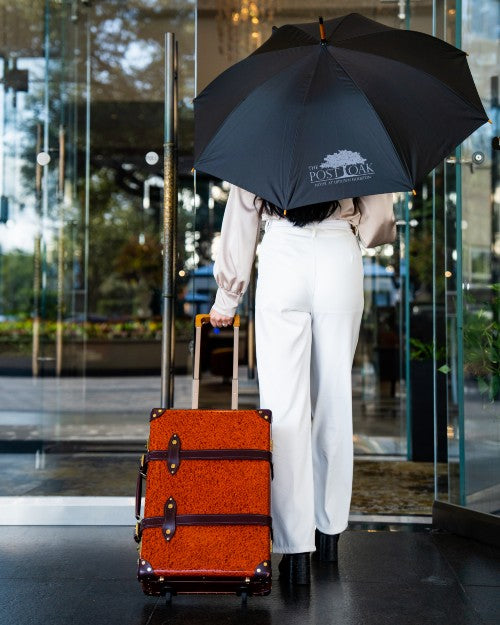 Woman walking into The Post Oak Hotel while carrying The Post Oak Umbrella and luggage.