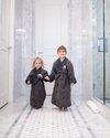 Two small children wearing The Post Oak Kids Robe in charcoal while standing in white hotel bathroom.