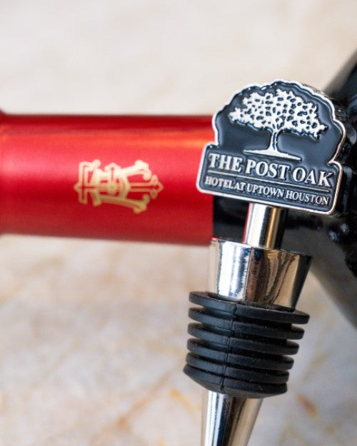 Silver wine stopper with The Post Oak logo at the top in front of red wine bottle. 