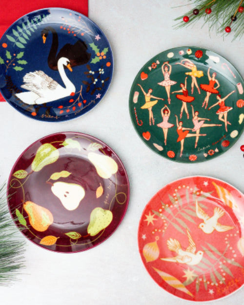 4 multicolored plates with different Christmas designs laid on white surface with tree needles in corners.