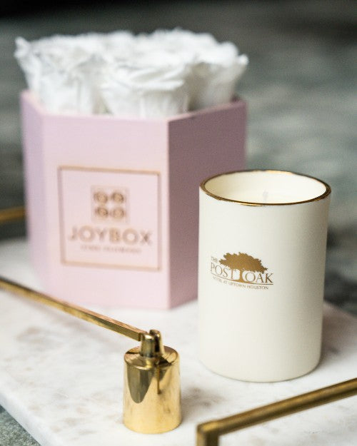 The Post Oak White Oak Candle on white marble table next to gift box wrapping.