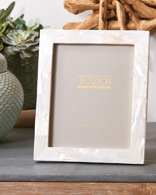 White mother of pearl photo frame placed on grey table surrounded with vibrant plants.