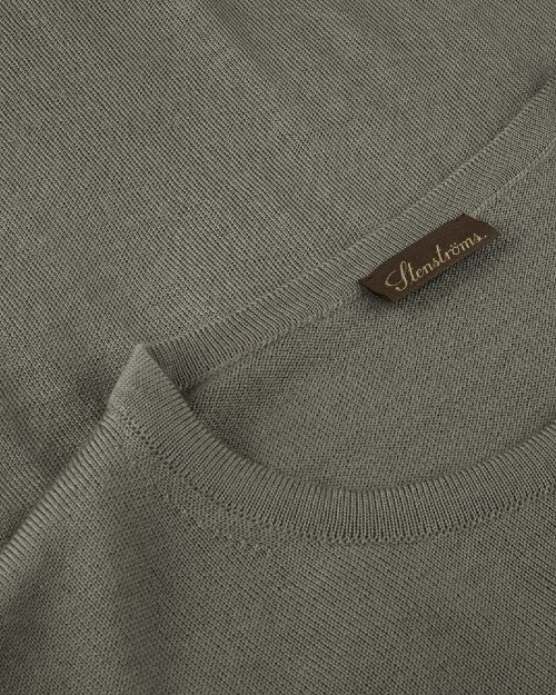 A close-up view of a folded green shirt's crewneck neckline, with a rectangular brown tag reading "STENSTRӦMS". 