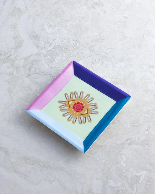White plate with multicolored edges and evil eye in center placed on marble table.