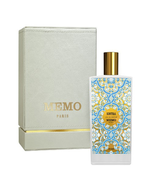 A rectangular white perfume bottle with a golden cap and a rectangular label featuring an intricate blue and yellow mosaic pattern, with a round gold label reading 'SINTRA Eau de Parfum MEMO Paris.' The bottle is placed against a light-colored box with a gold stripe at the bottom and "MEMO PARIS" printed in gold above it. The background is plain white.