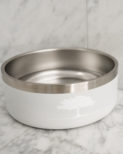 A white dog bowl with a silver rim and a white tree design on the front. The background is a marble surface.
