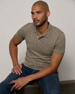 Model wearing green/tan polo with jeans while sitting on bench.